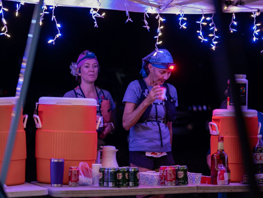 Nighttime aid station with string lights and volunteers serving runners behind orange coolers.