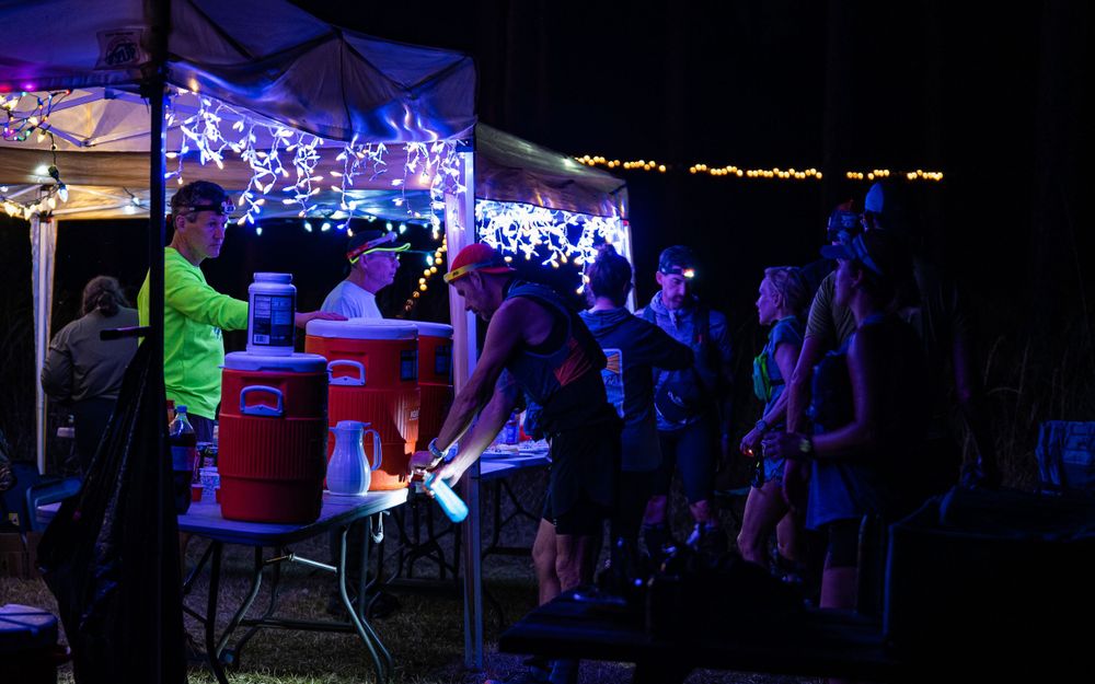 Purple-lit outdoor race aid station at night with volunteers and runners gathering.