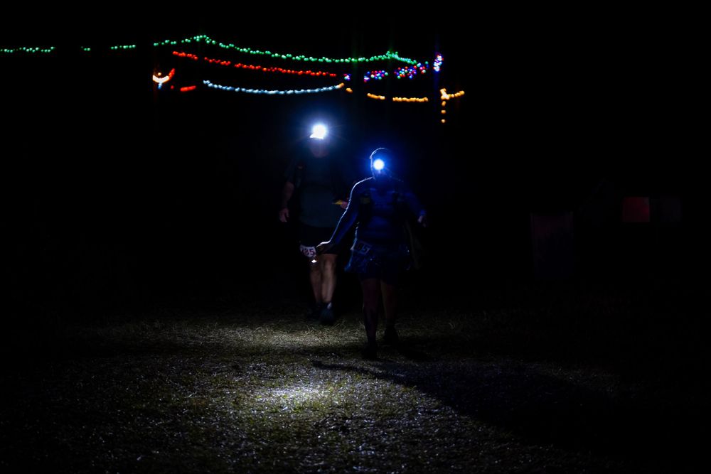Runners illuminated by headlamps passing under colorful string lights on dark trail.