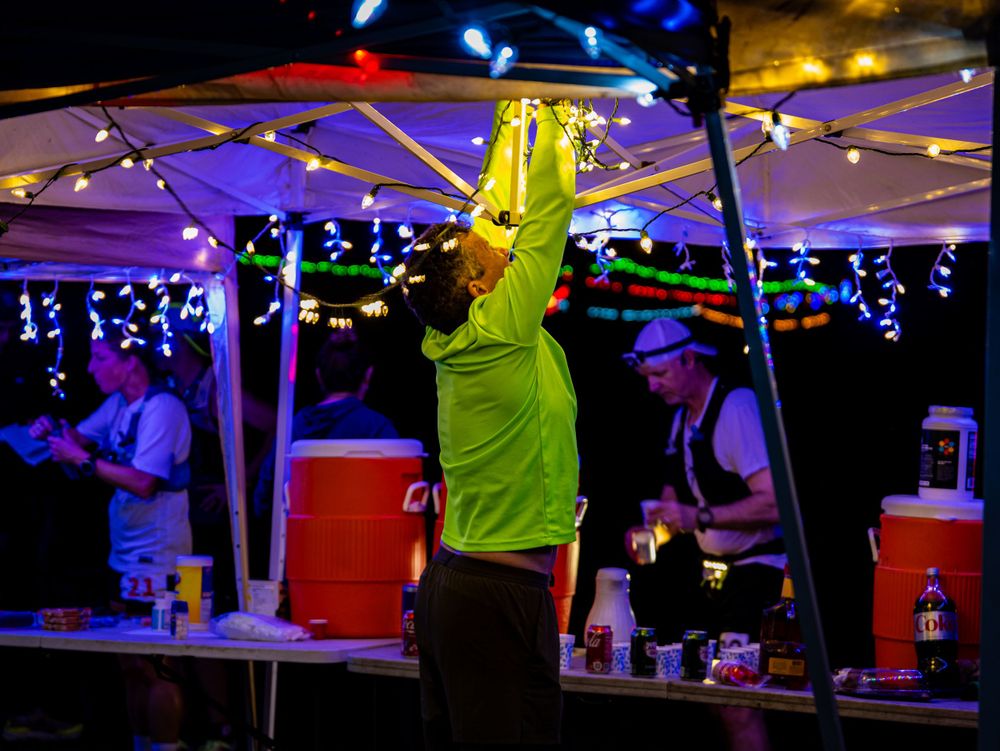 Aid station volunteer in bright yellow shirt serving runners under purple string lights.