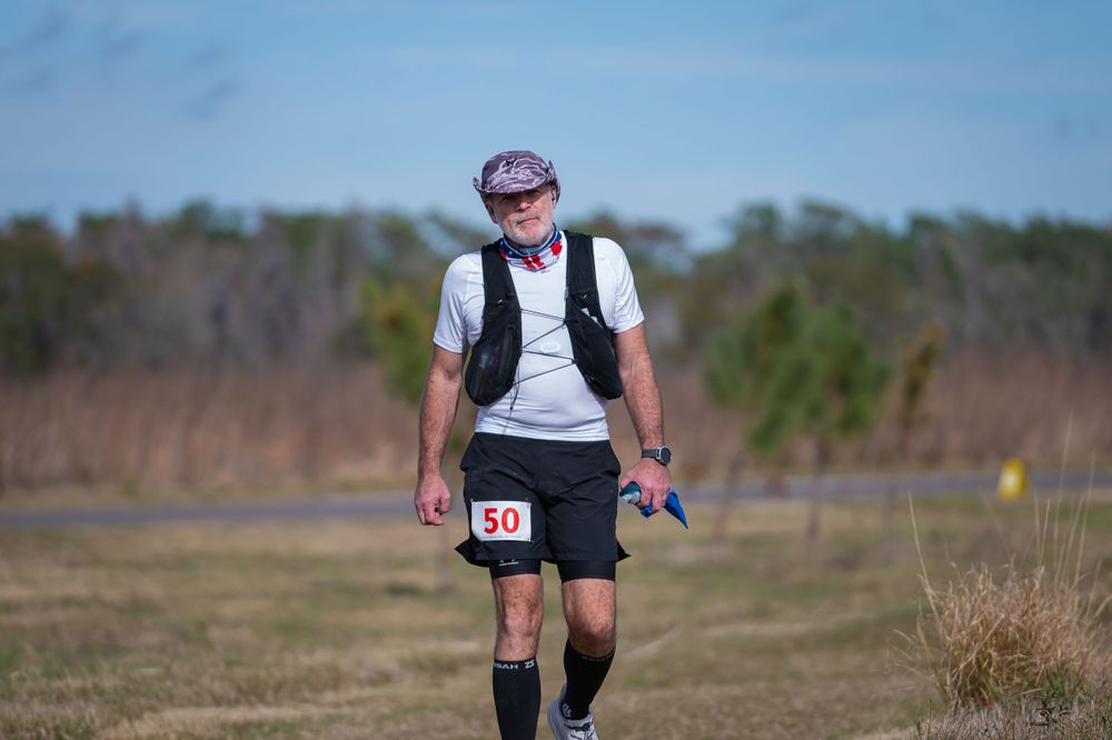 Runner wearing backpack and hat traversing trail through open field under blue sky.