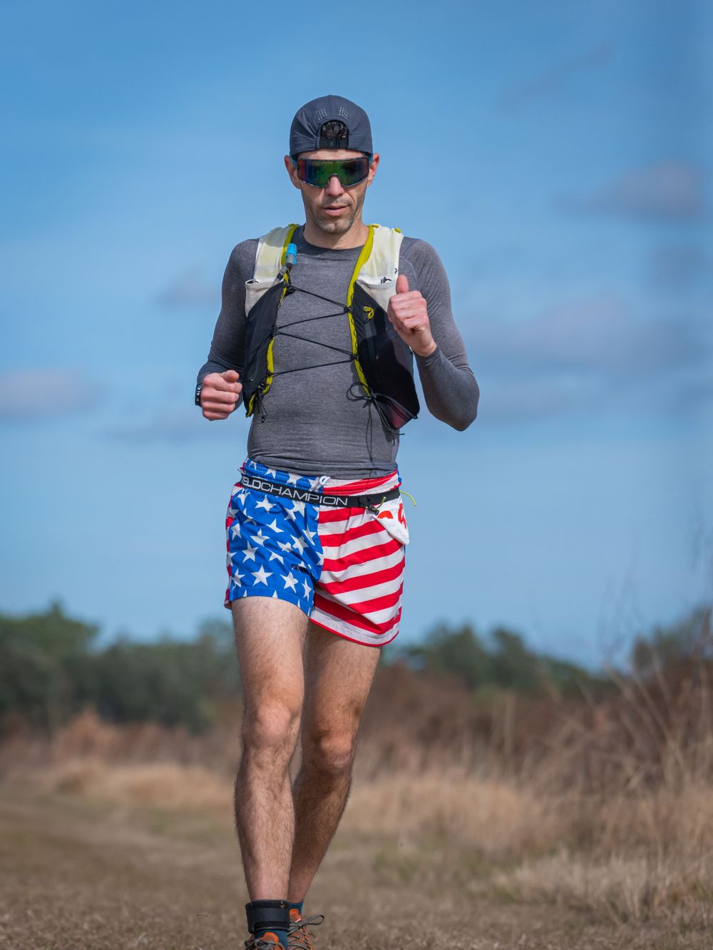 Runner wearing American flag shorts and hydration pack running on trail under blue sky.