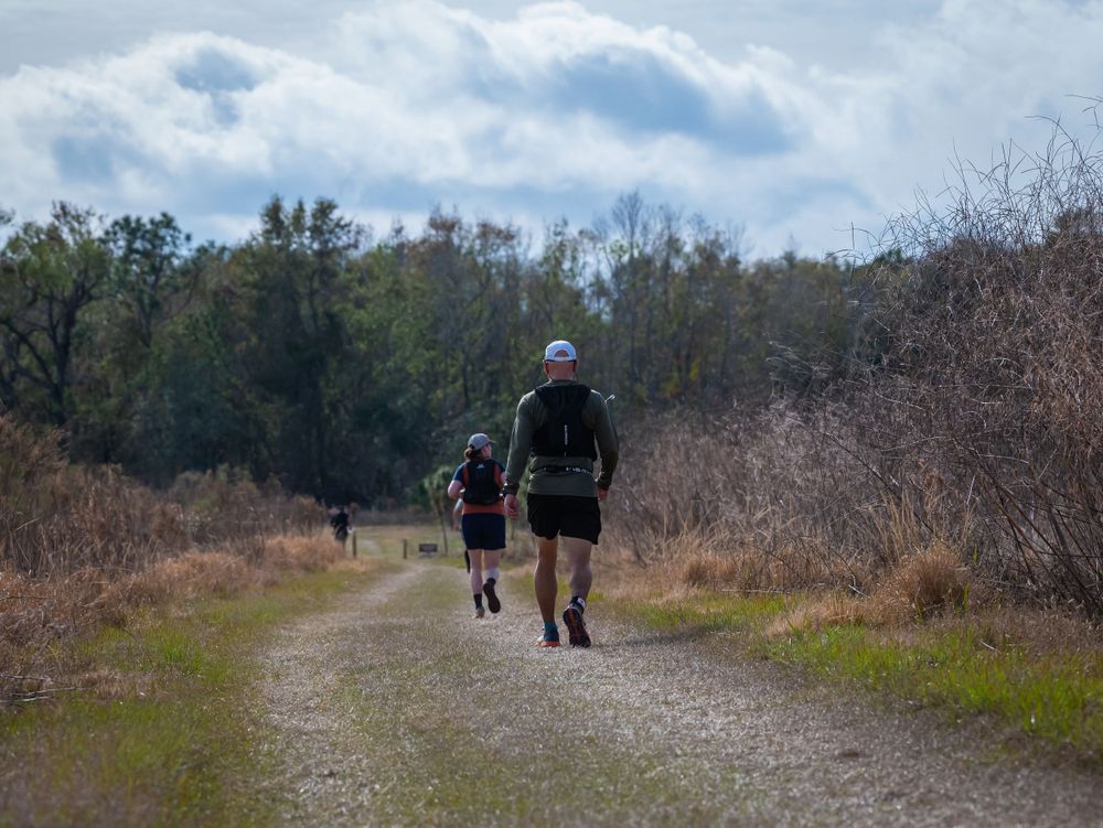 Trail runners on dirt path through brown winter brush under cloudy skies.