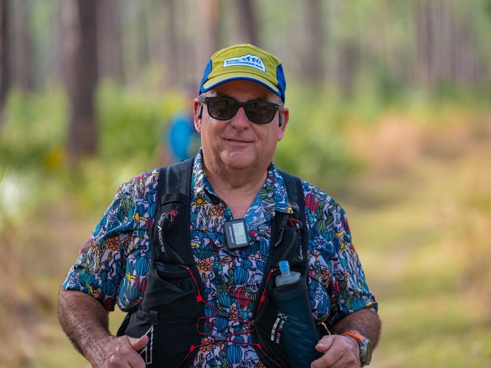 Trail runner wearing floral shirt and backpack on wooded path.