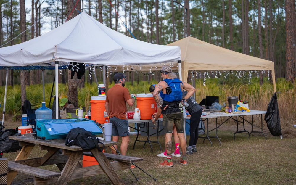 Trail running aid station with white canopy tents and volunteers helping runners.