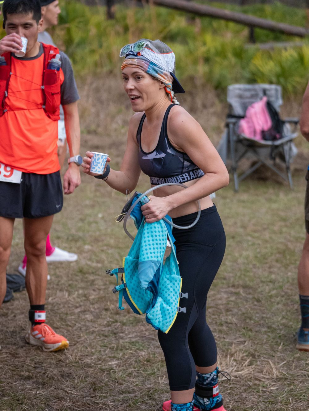 Trail runner with headband holding drinks and gear at outdoor race event.