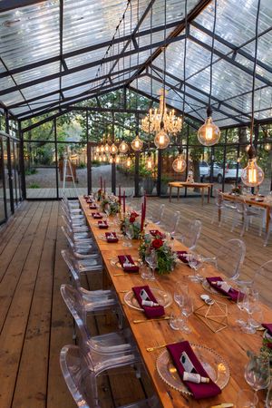 Elegant reception tables with burgundy accents and ghost chairs under hanging glass pendant lights.