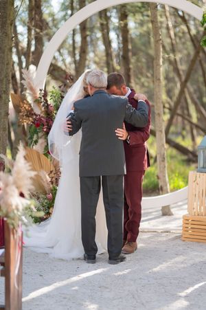Touching wedding ceremony moment under a white arch decorated with burgundy and white flowers.