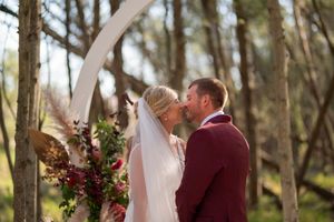 A romantic outdoor wedding ceremony features a burgundy suit and white dress against a wooden arch in a forest setting.