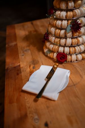 Macaron tower display with red roses and serving utensils on wooden table.