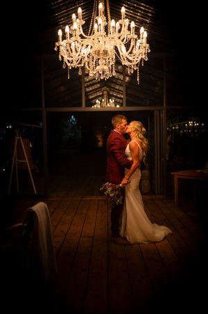 A romantic wedding moment under an elegant crystal chandelier in a rustic wooden venue at night.