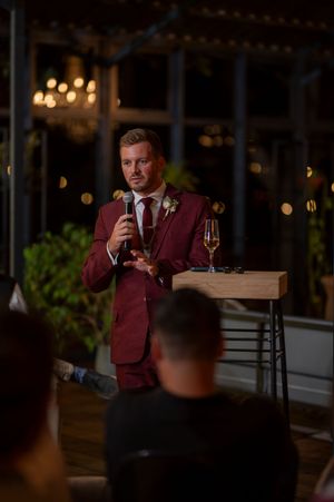 Wedding guest in burgundy suit delivering toast with microphone at evening reception.