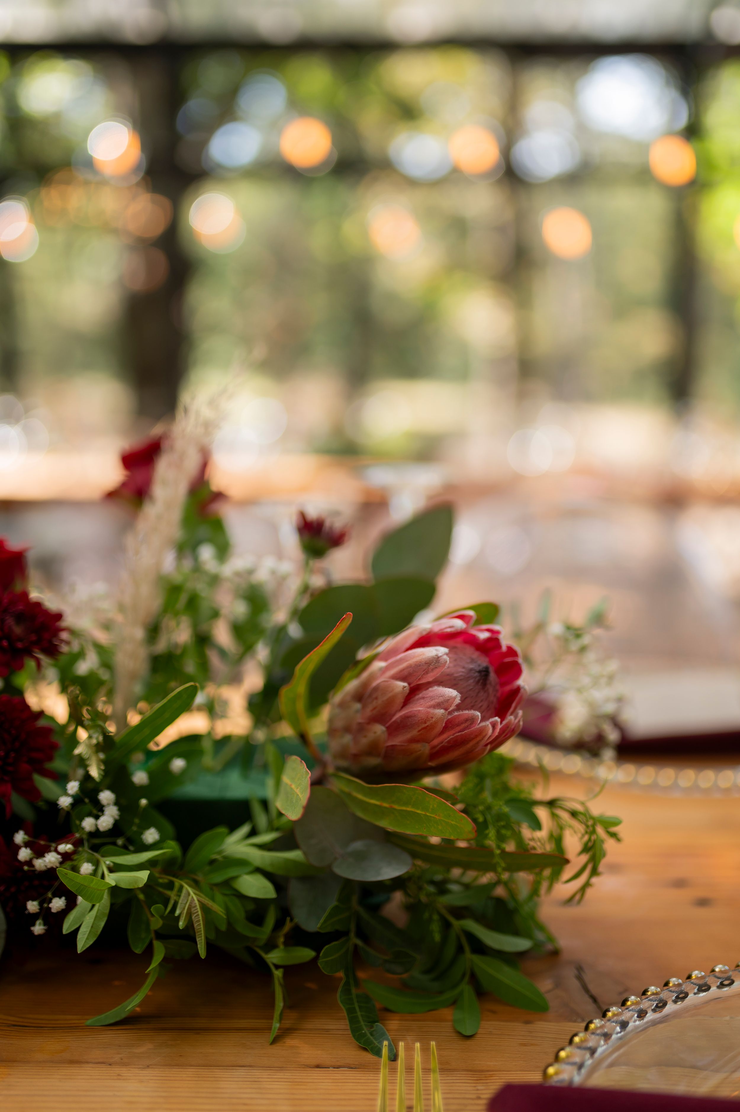 A romantic floral centerpiece with protea and greenery on a wooden table with bokeh lights.