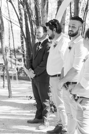 Three groomsmen stand in line at an outdoor forest wedding ceremony wearing different formal attire.