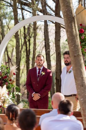 A wedding ceremony in a wooded setting with a white arch decorated with red flowers.