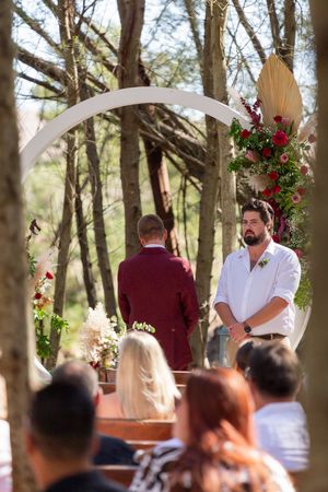 Wedding ceremony in progress under white arch with floral decorations in a forest setting.