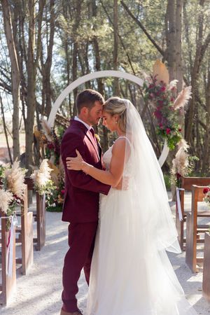 Newlyweds share intimate moment under white floral arch during forest wedding ceremony.