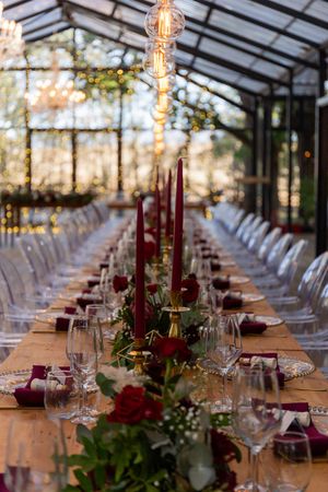 Elegant wedding reception table setup with burgundy accents and hanging Edison bulbs in glass venue.