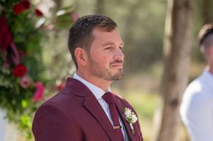 A wedding ceremony participant in burgundy suit standing at forest venue with floral decorations.
