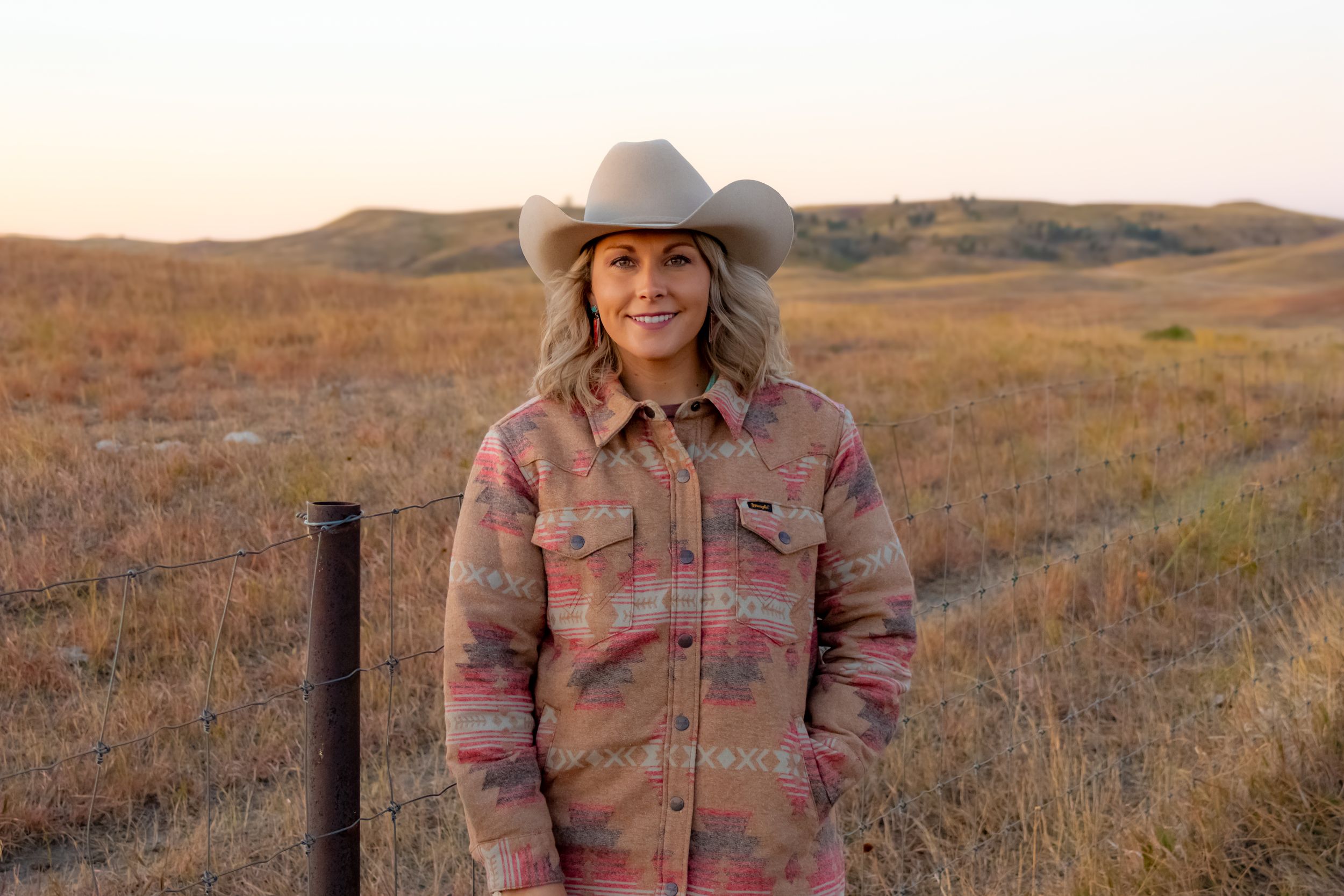 Person wearing a cowboy hat and plaid jacket standing in a rustic field at sunset.