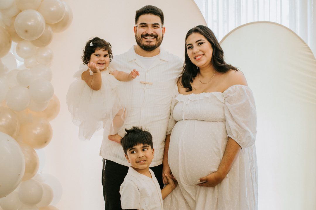 A family poses at a baby shower celebration with white balloons and decorations in the background.
