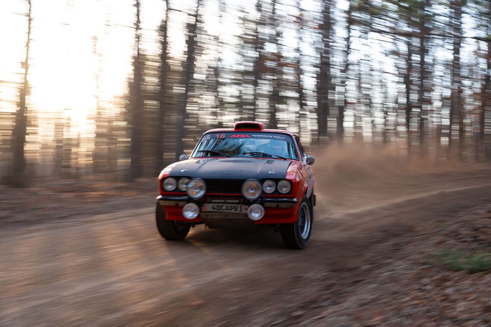 Rally car kicking up dust while drifting through forested dirt road.