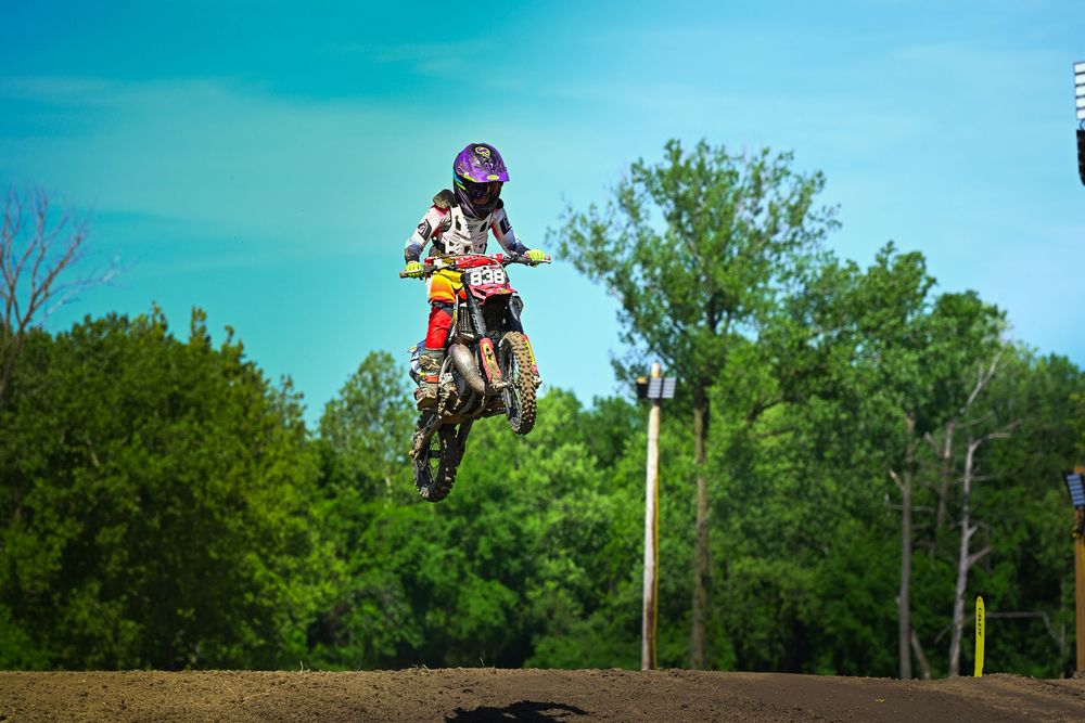 Motocross rider in purple helmet catches air on red dirt bike at outdoor track with green trees and blue sky.