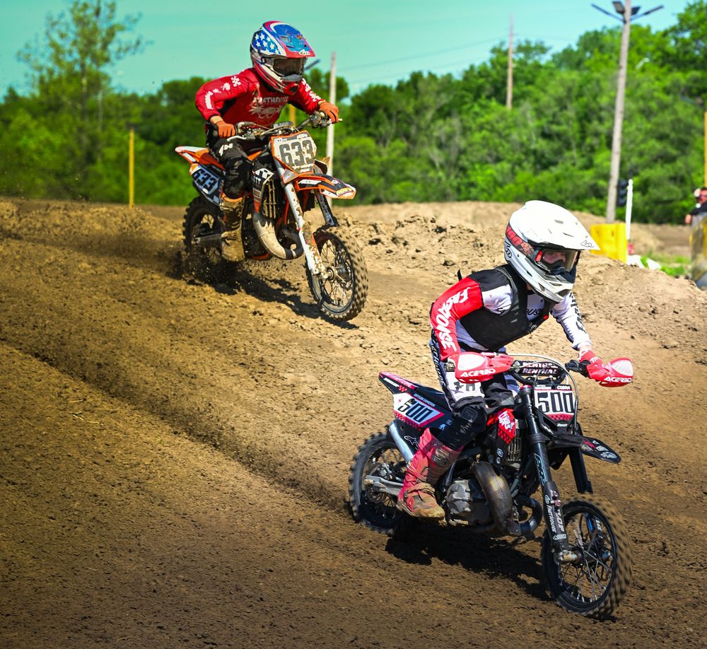 Two motocross riders race on a dirt track, wearing red gear, on numbered bikes 633 and 500 during an outdoor competition.