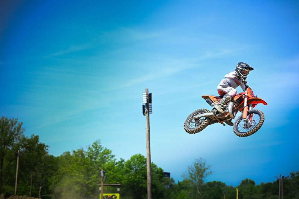 Motocross rider performing a high jump trick on an orange dirt bike against a clear blue sky.