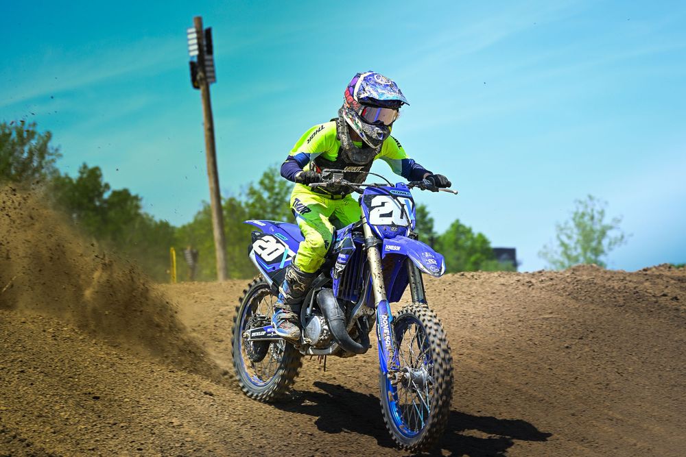 Young motocross rider in green gear racing blue dirt bike #21 on a sandy track.