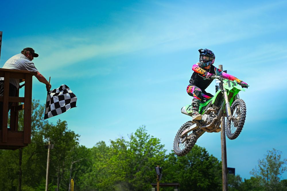 Motocross rider jumps green dirt bike near checkered flag waver on platform against blue sky.