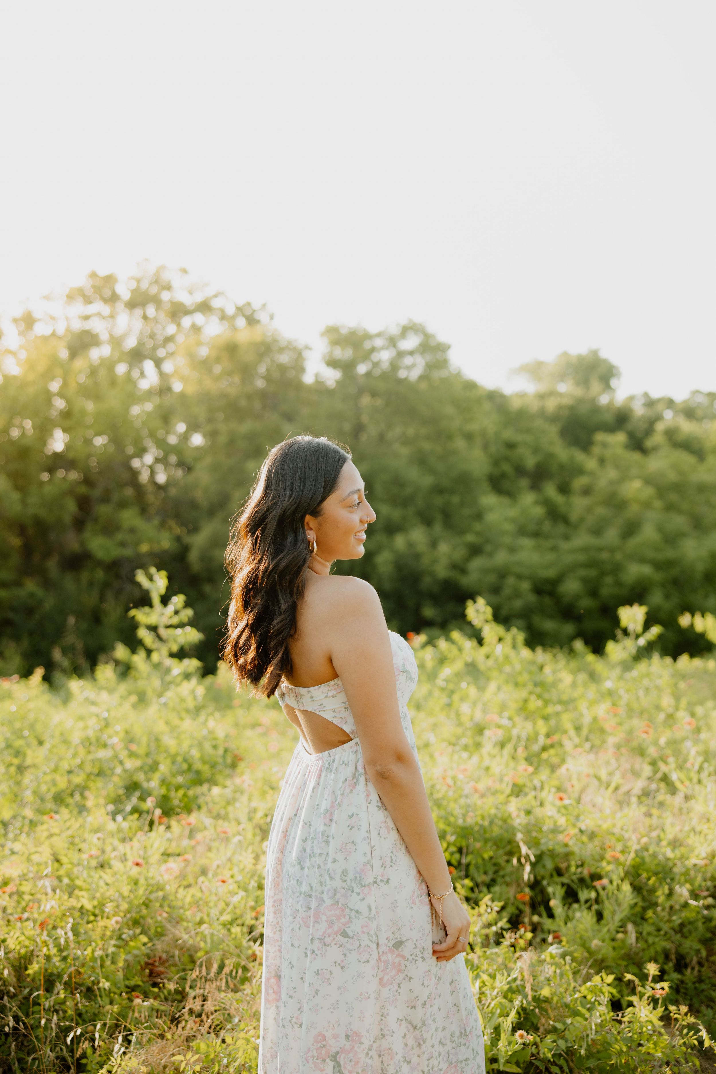 Person in white floral sundress stands in golden sunlit meadow during sunset outdoors in nature.