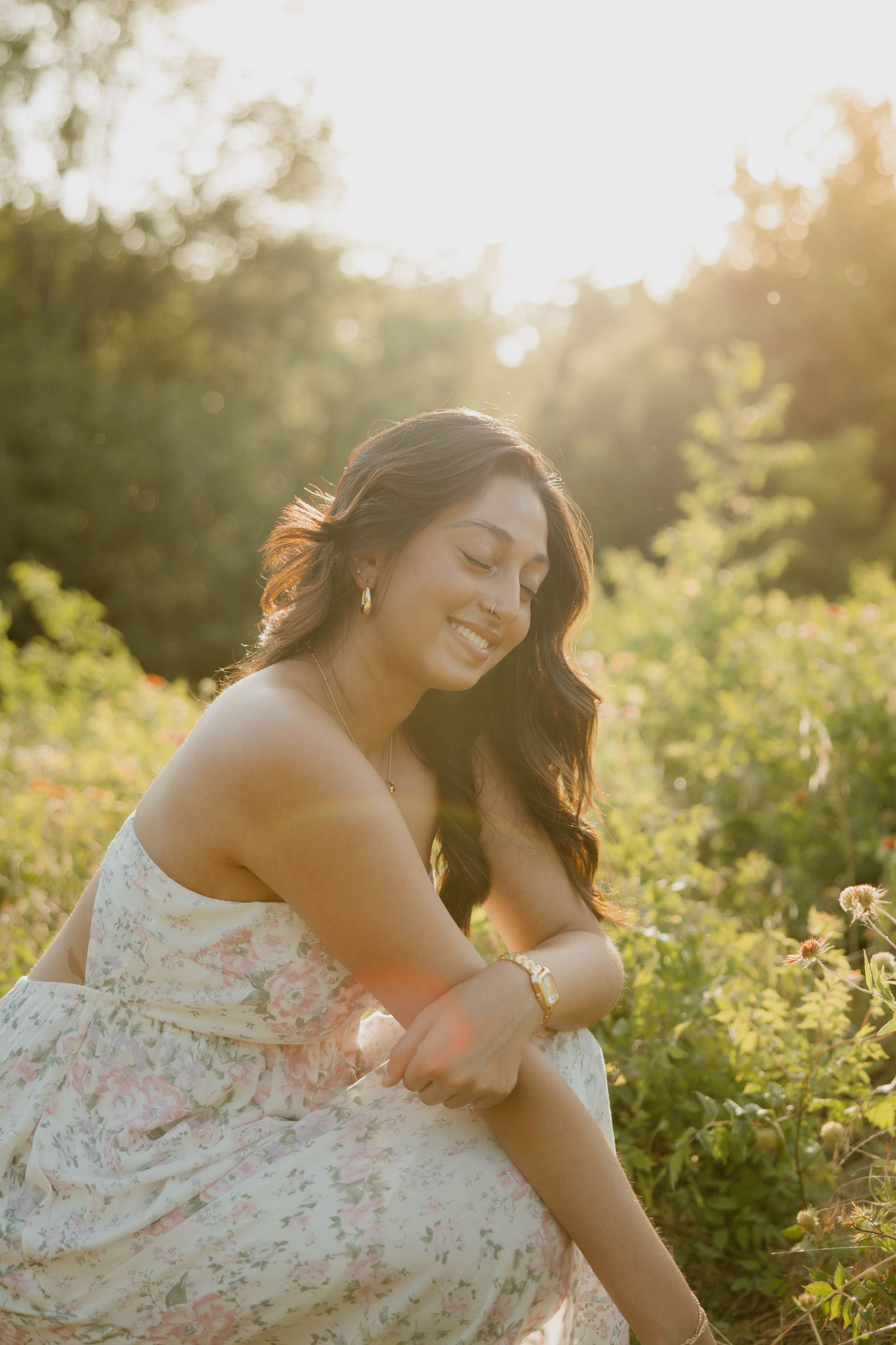 A person in a floral dress sits peacefully in a sunlit meadow during golden hour, surrounded by wildflowers and greenery.