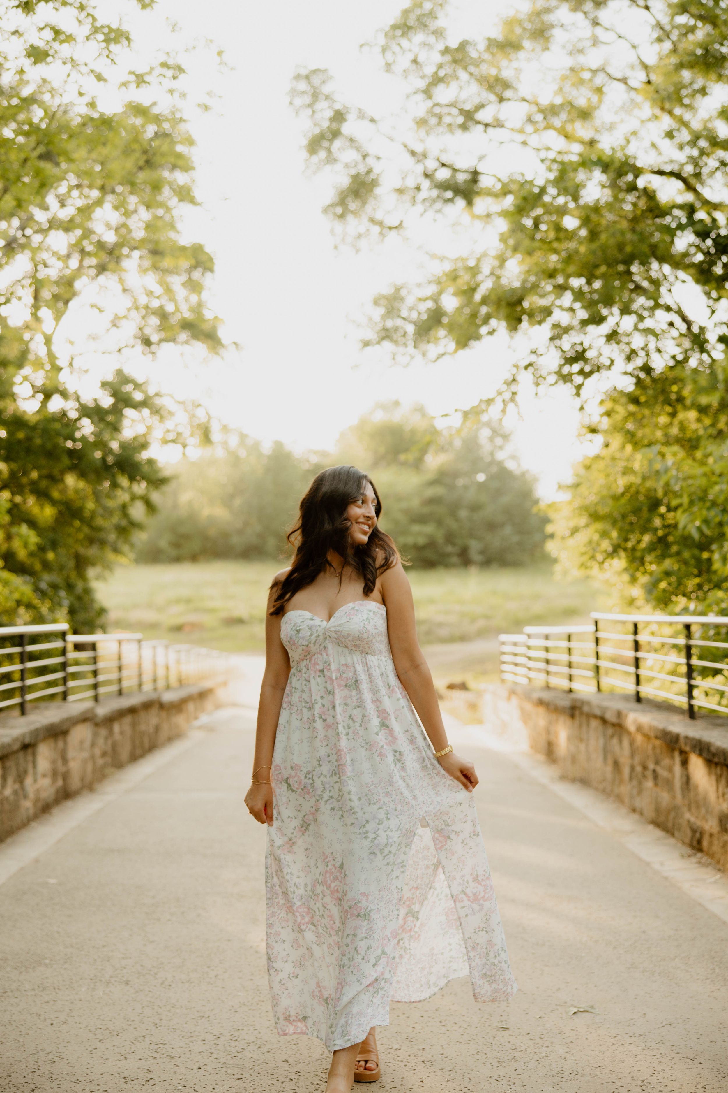 A white floral sundress flows gracefully along a tree-lined path with wooden fences at golden hour.