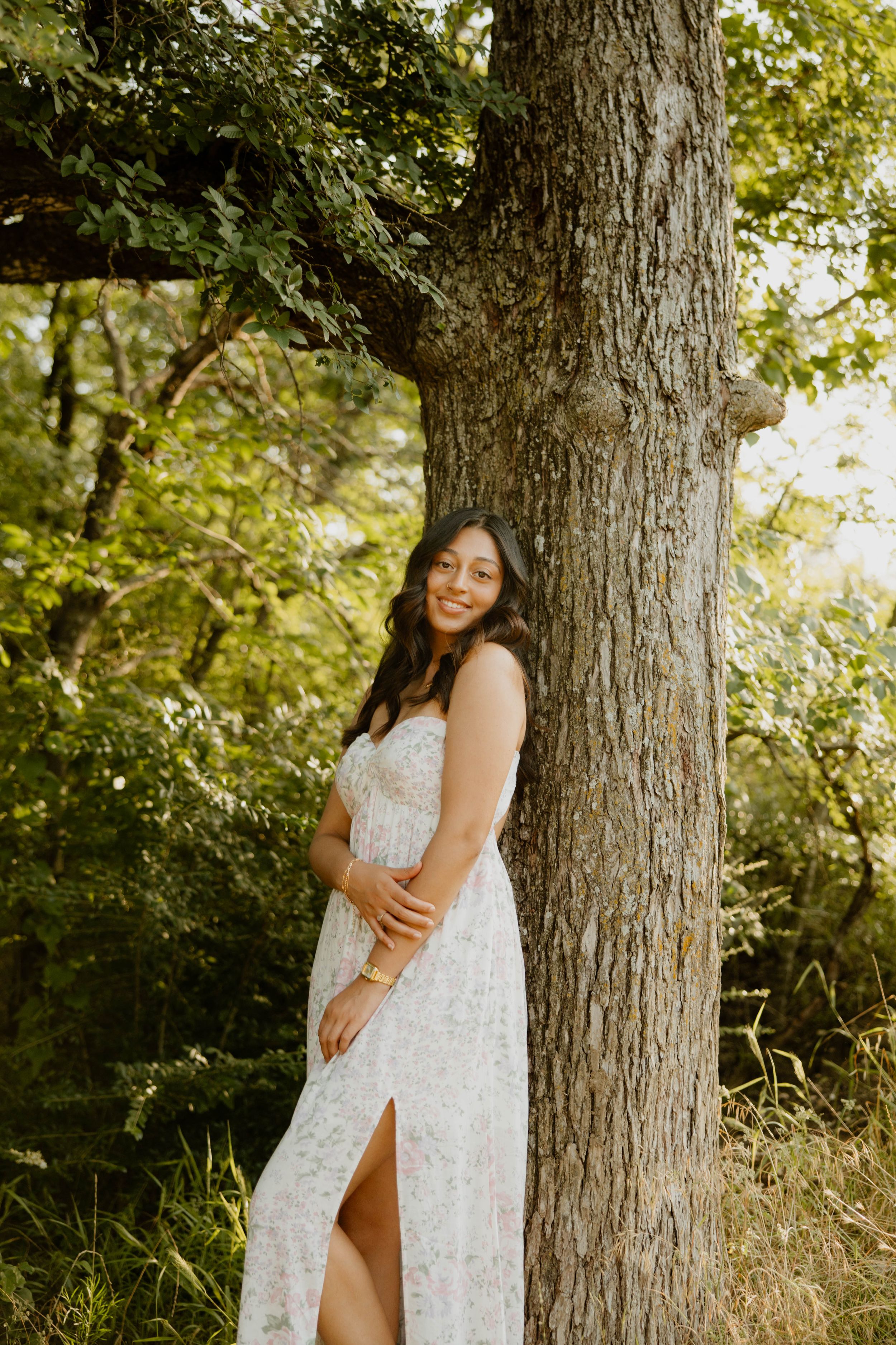 A woman in a white lace dress poses next to a large tree trunk in a sunny outdoor setting.