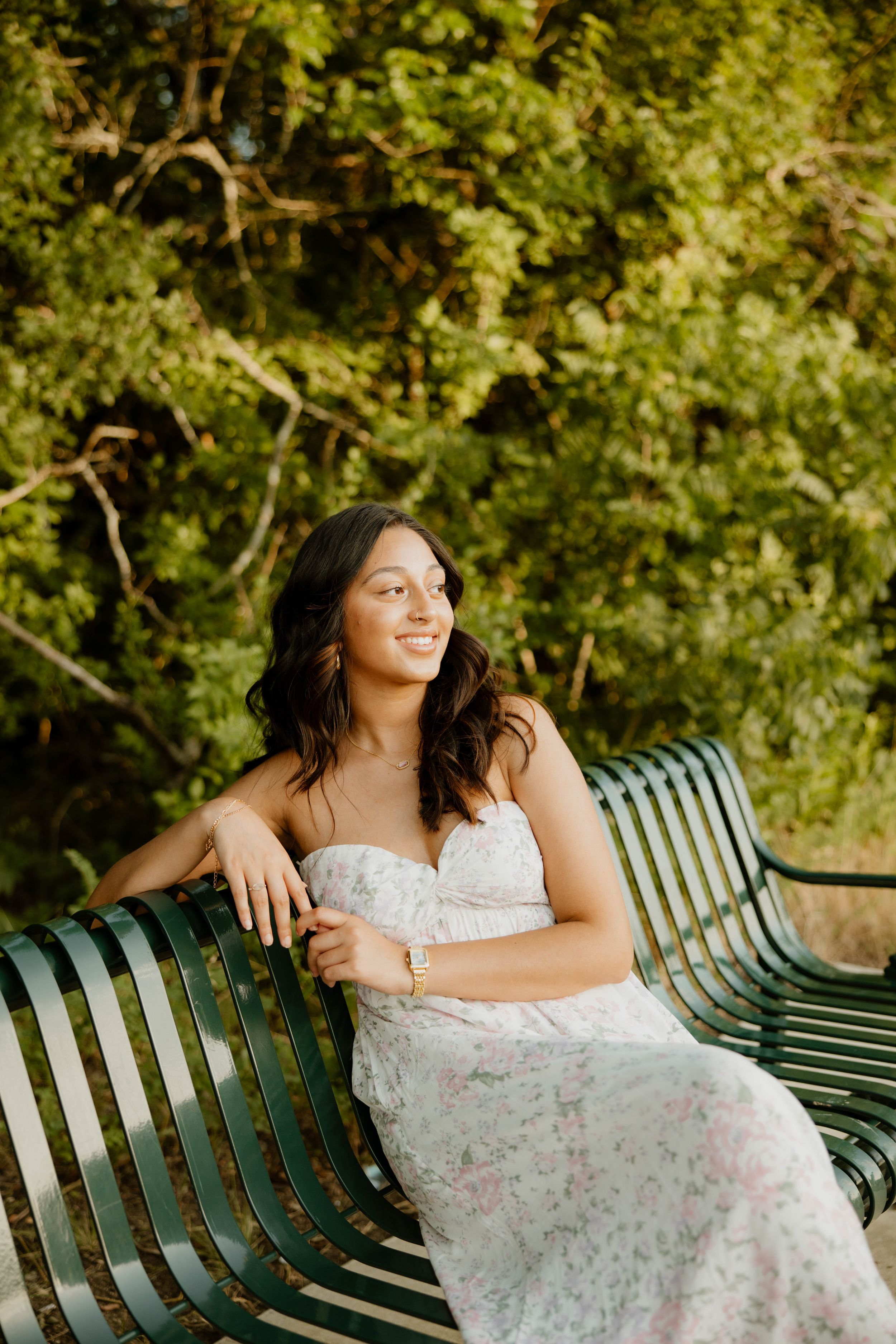 A person wearing a delicate white floral dress sits on a green park bench surrounded by lush greenery on a sunny day.