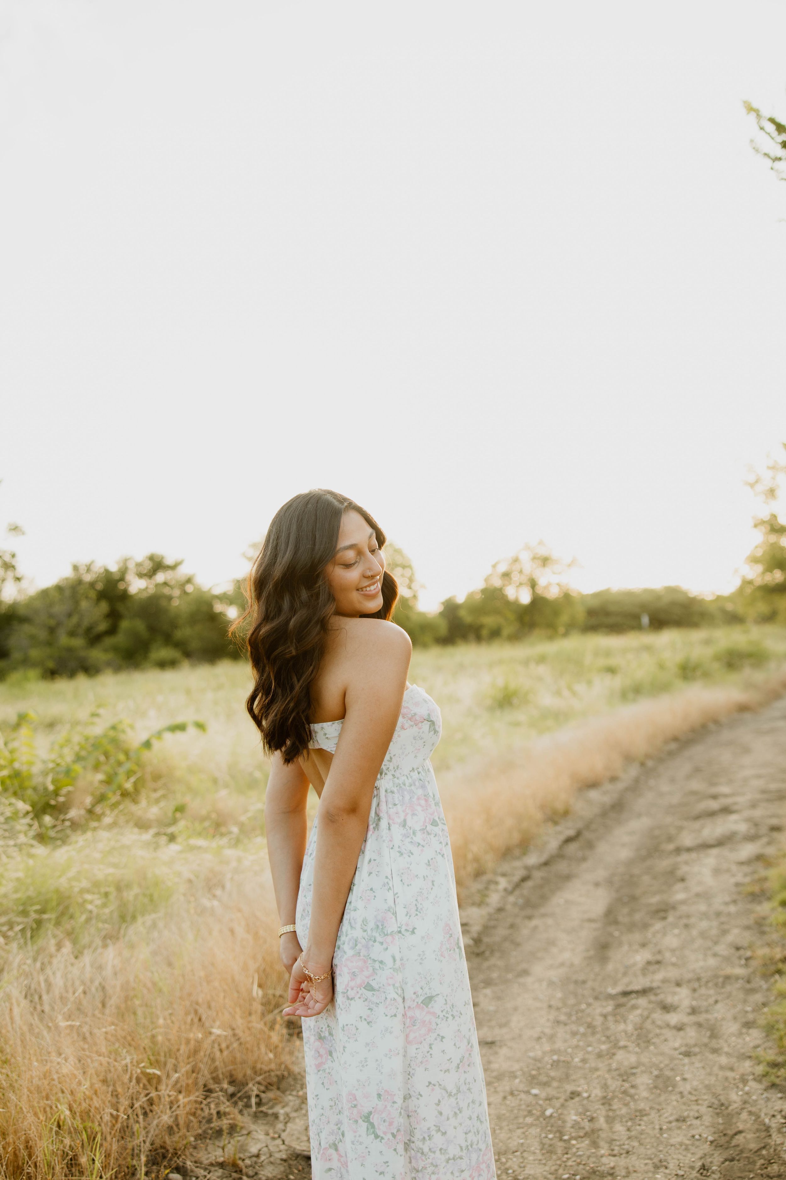 A person in a floral maxi dress stands along a dirt path at golden hour in a natural outdoor setting.