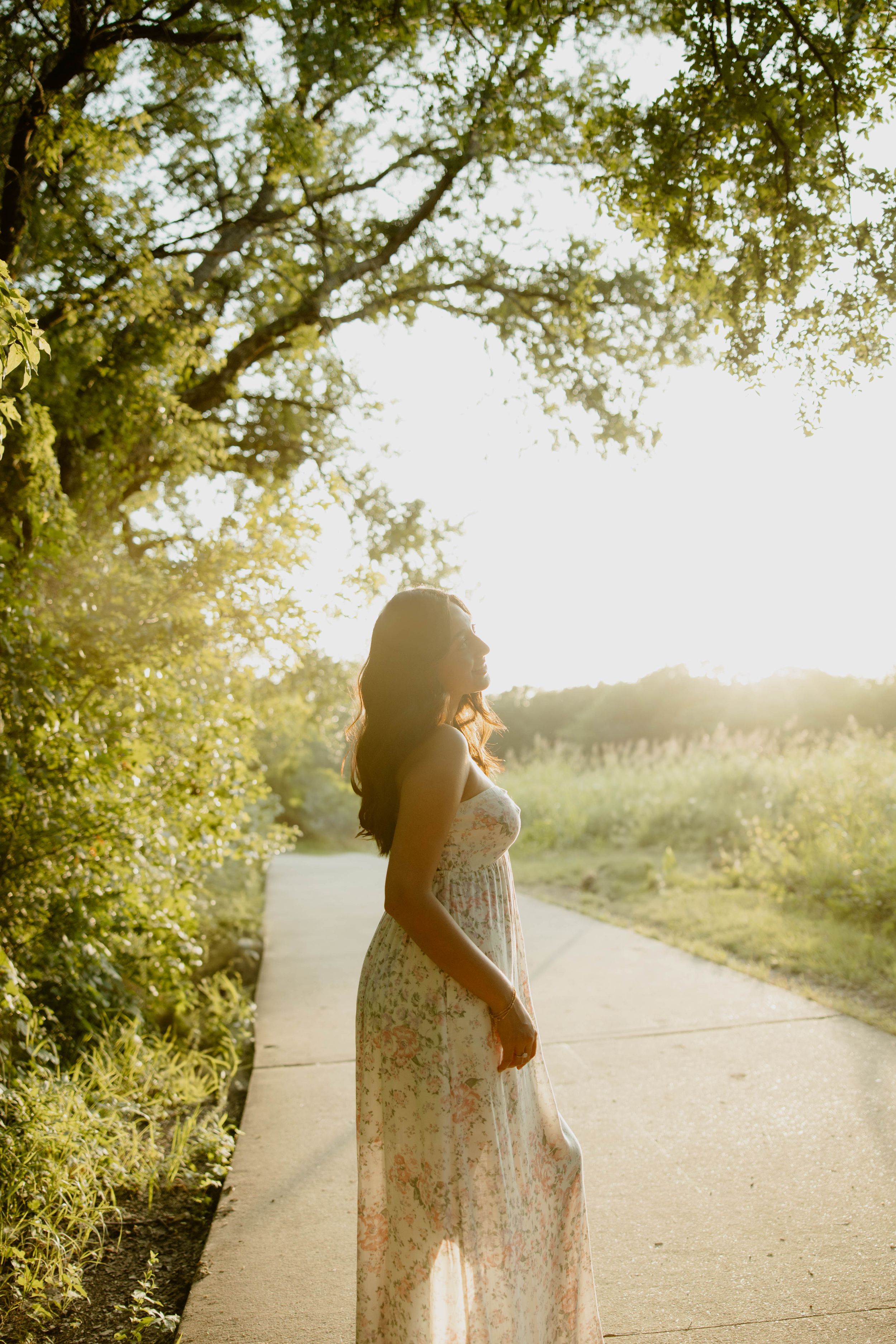 Sun-drenched silhouette of a person in a flowy lace dress standing on a paved path amidst green foliage during golden hour.