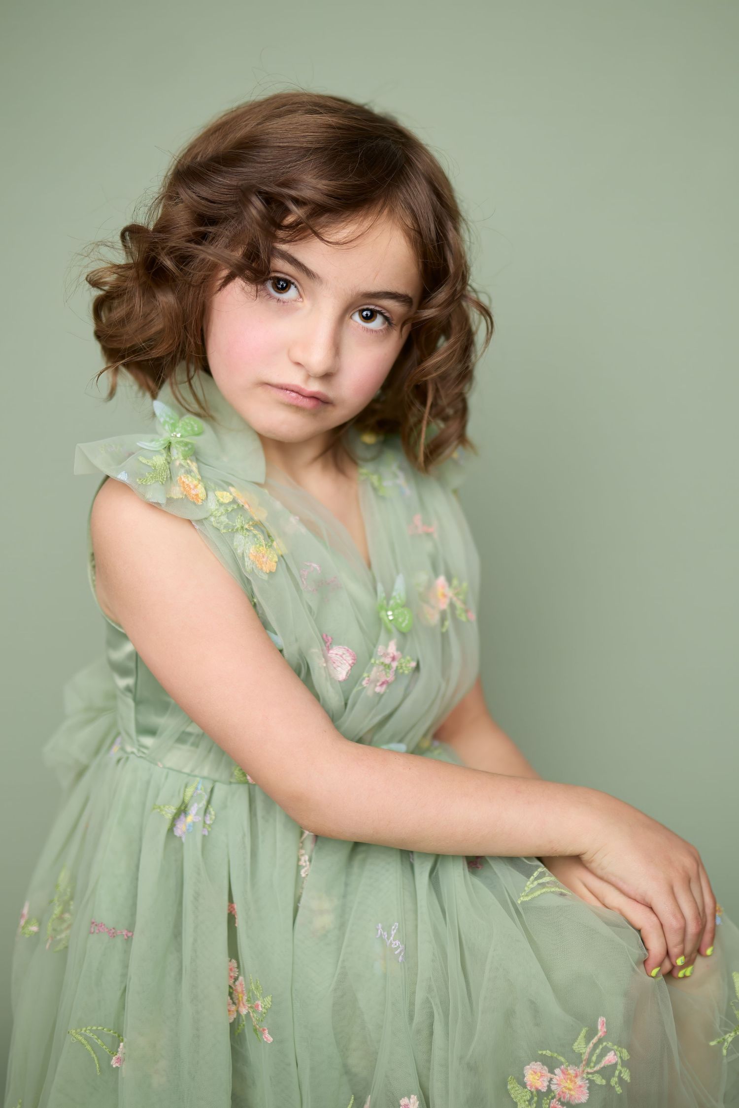 Young child wearing a mint green floral dress with delicate embroidered details poses against a light backdrop in a portrait style.