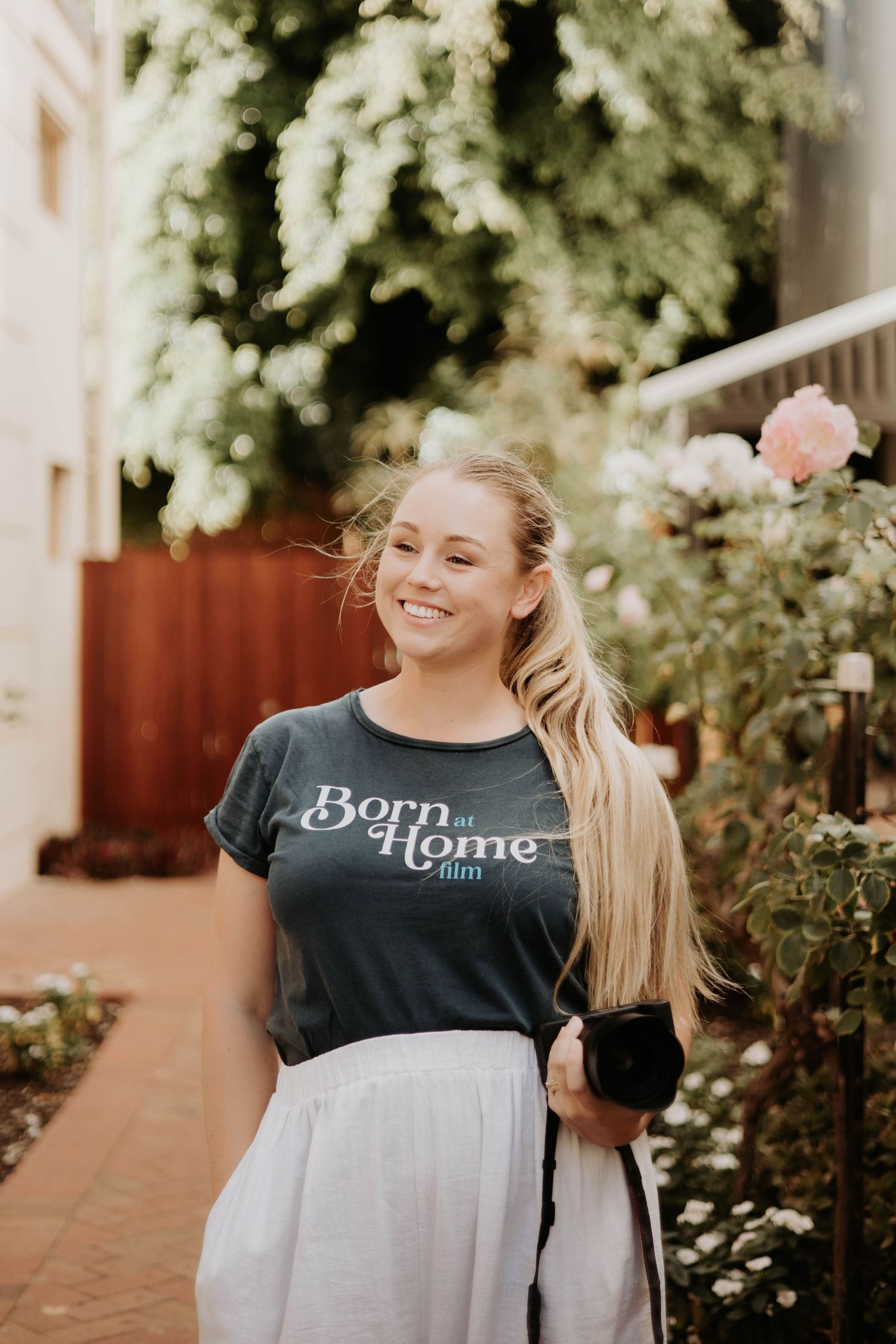 A woman in a grey "Born at Home" t-shirt and white skirt stands smiling in a garden pathway with flowers and greenery.