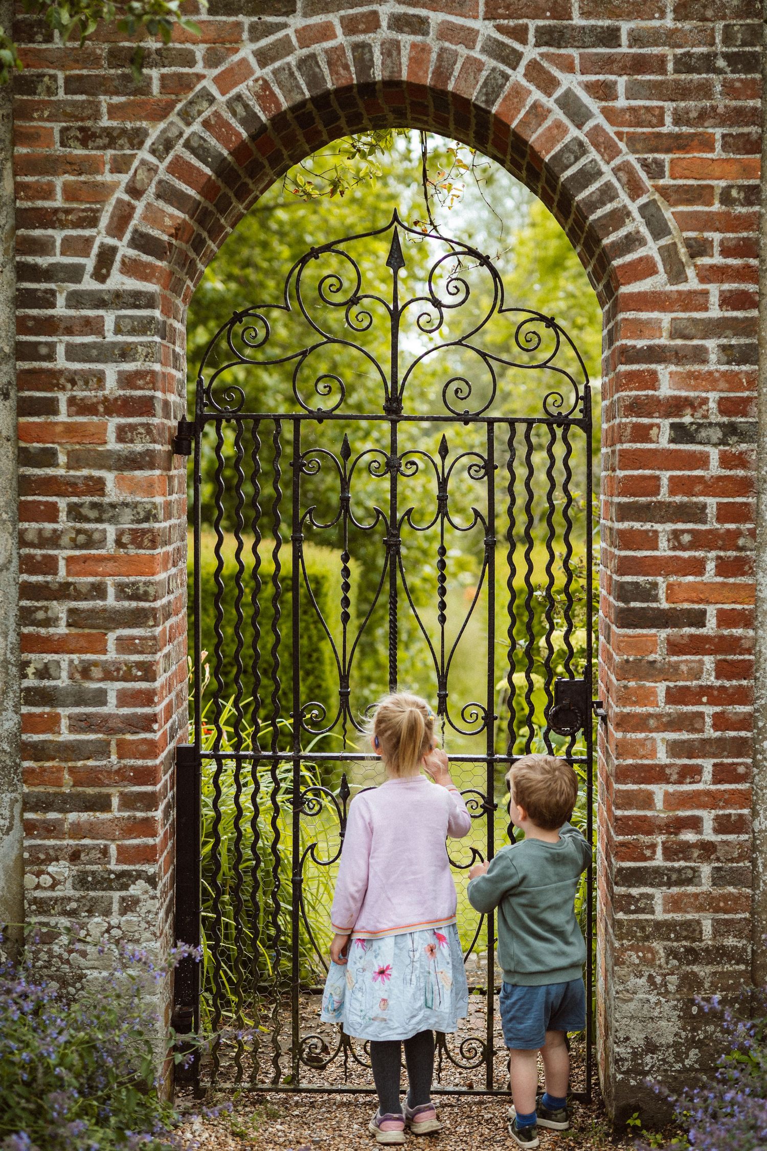 Two small children peer through an ornate wrought iron garden gate framed by a brick archway.