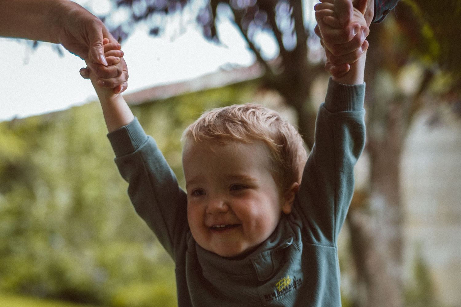A joyful child with raised arms being lifted up outdoors on a sunny day.