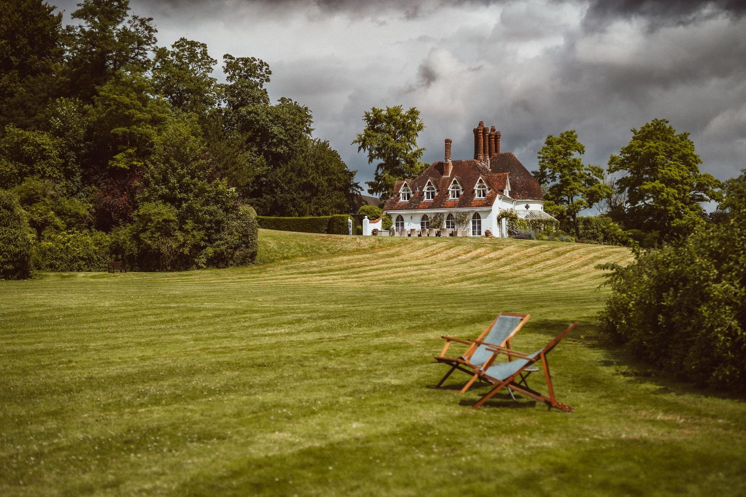 Deck chair sits on manicured lawn overlooking English country house with dark storm clouds gathering above.