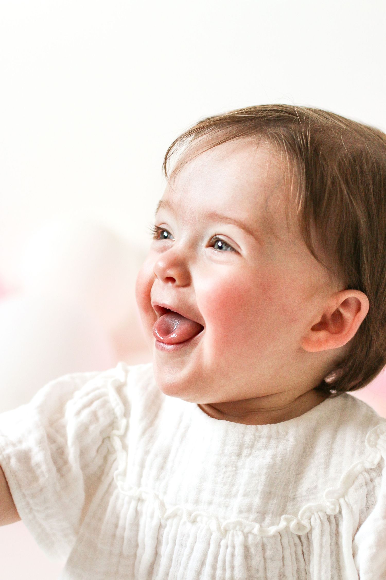Joyful baby wearing white dress smiles with rosy cheeks in bright natural light, during birthday shoot in Nottingham