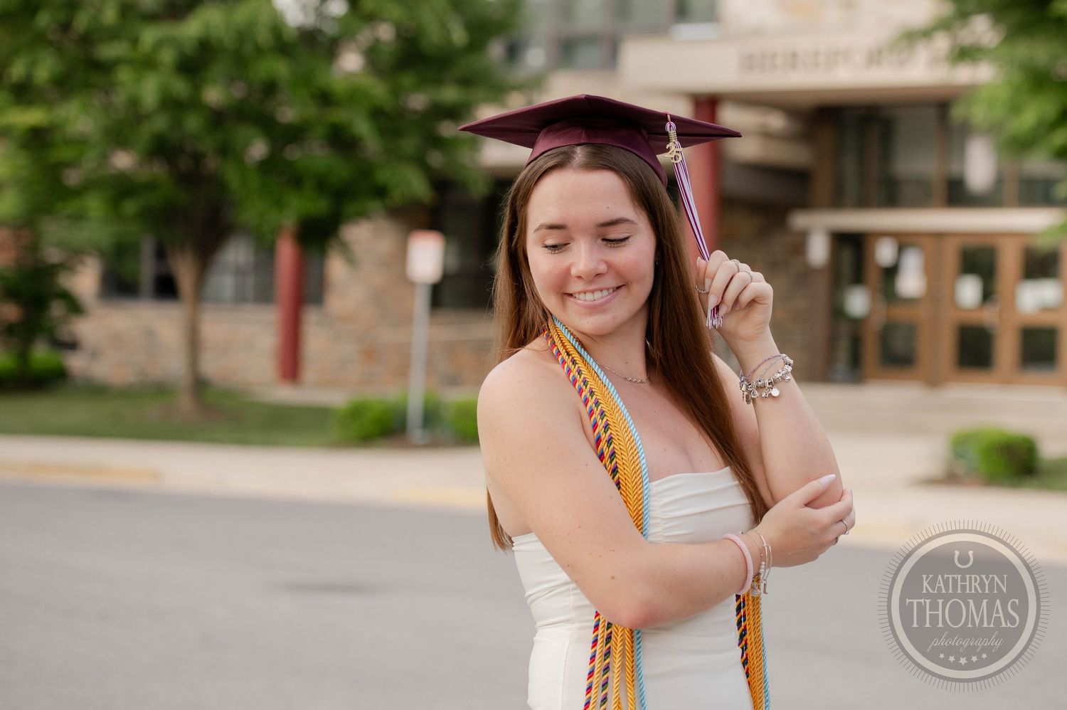 Graduate in white dress wearing burgundy cap and gold honor cords poses outdoors during commencement ceremony.