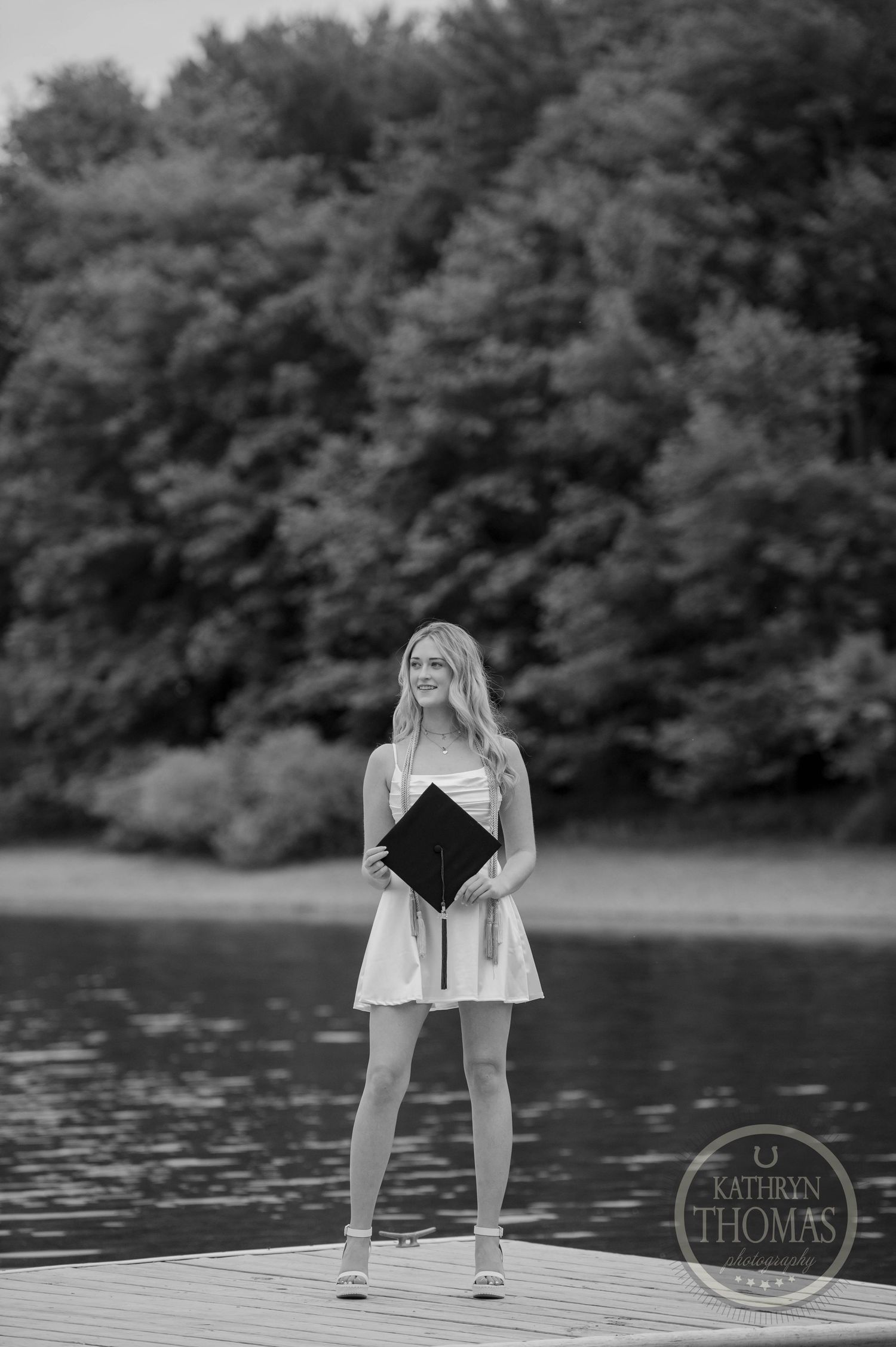 A graduate stands on a dock by a lake holding their diploma in a black and white portrait photo.