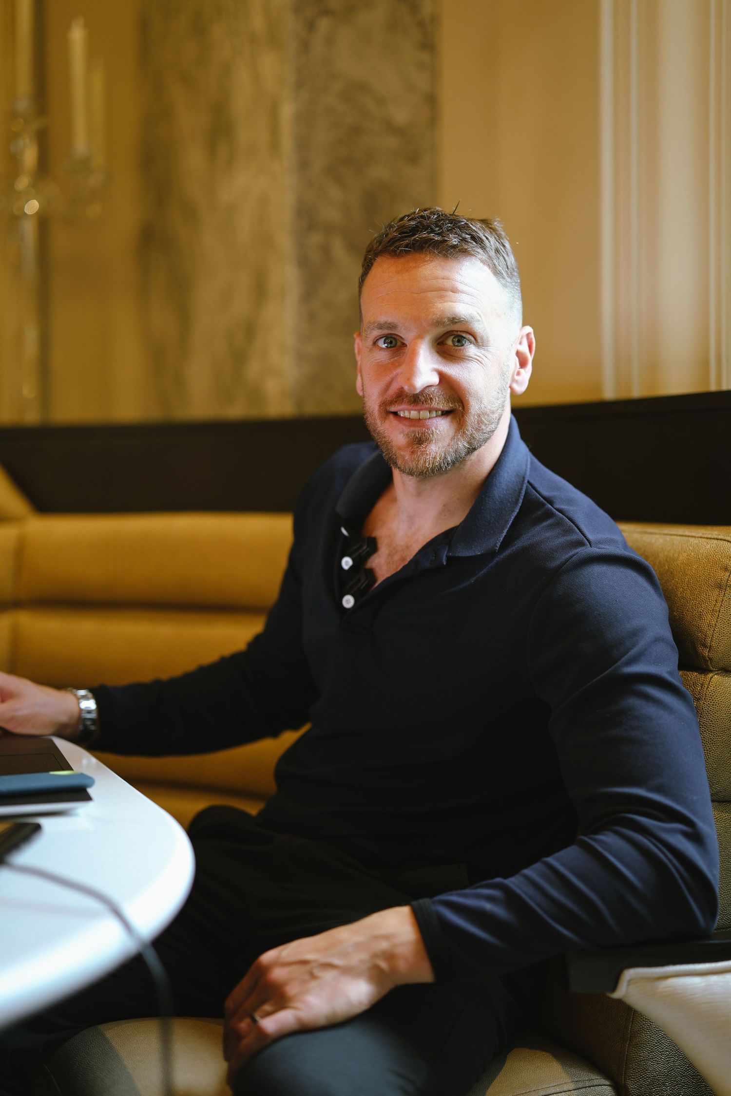 A man in a navy polo shirt sits casually on a yellow couch in an elegant room.