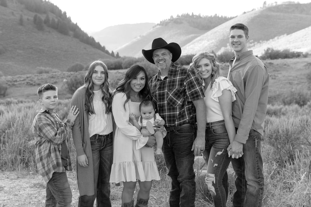 Family group poses together outdoors with rolling hills and mountains in black and white photograph.