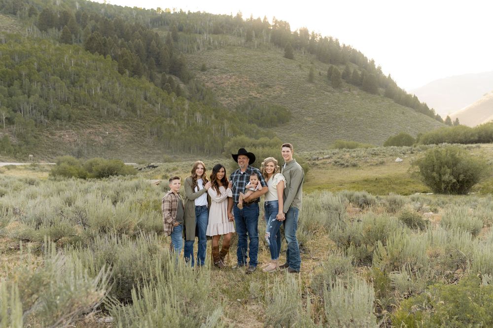 Family group stands together outdoors in rural mountain setting with sagebrush and hills in background.