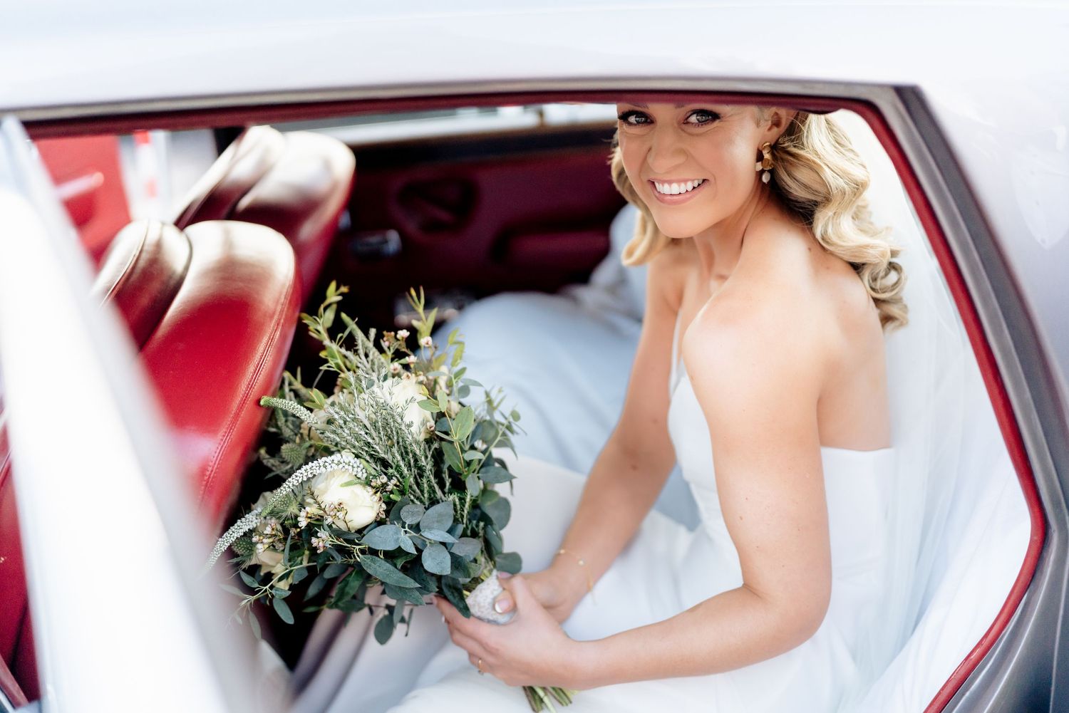 A bride sitting in a vintage red car holding a beautiful green and white wedding bouquet.